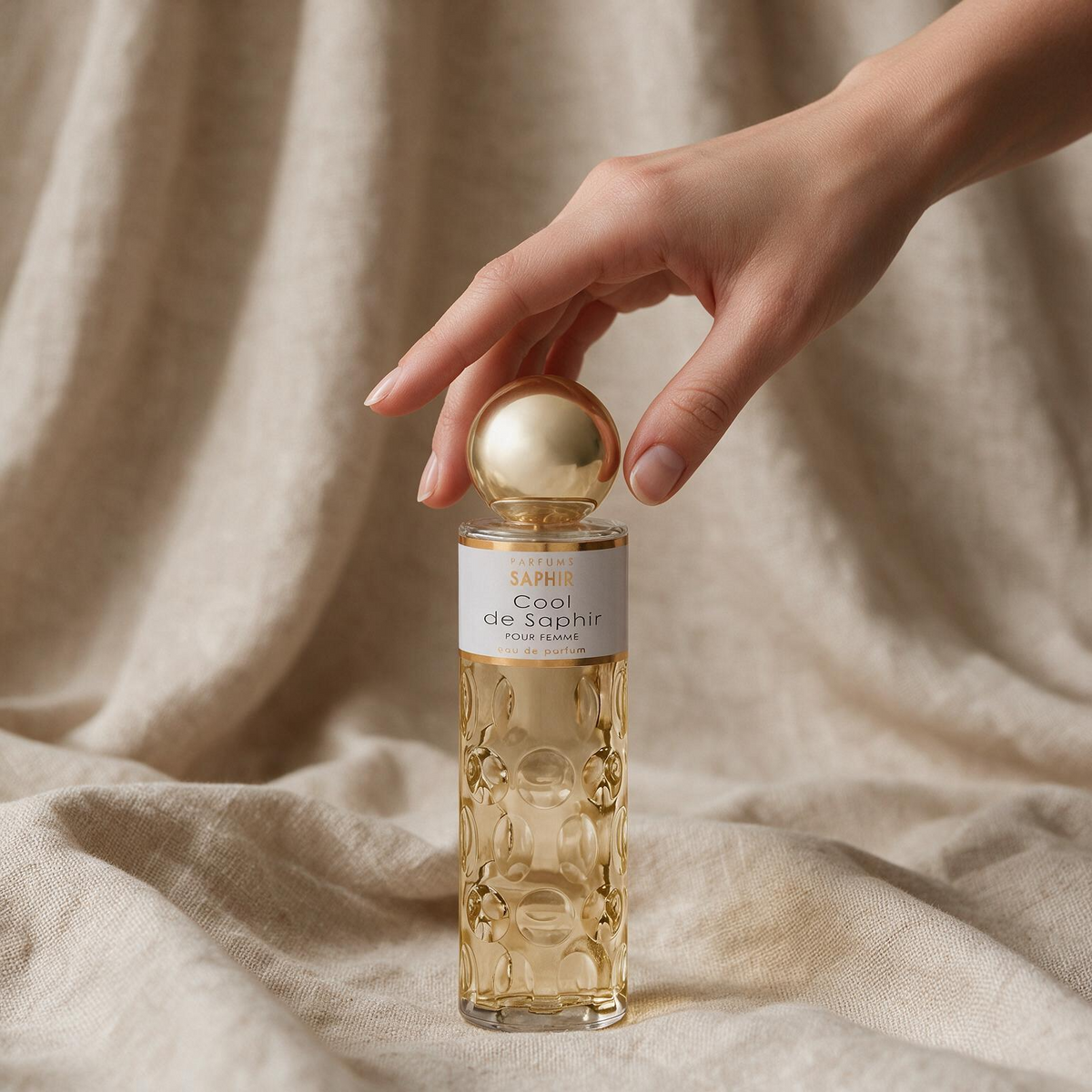 Hand reaching for Cool de Saphir perfume bottle on linen fabric backdrop